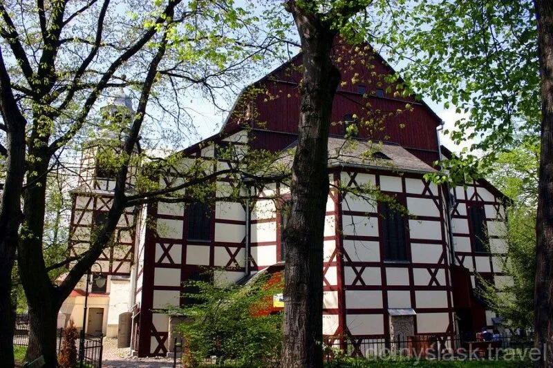 La façade à colombages de l'Église de la Paix à Jawor, avec ses panneaux blancs et ses poutres sombres, s'élève au milieu des arbres verts, partiellement masquée par des branches printanières.