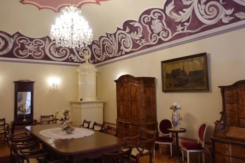 Oświetlone wnętrze pokoju w muzeum ze zdobionym sufitem obrazem na ścianie, drewnianą starą szafą i stołem w centralnej części pomieszczenia