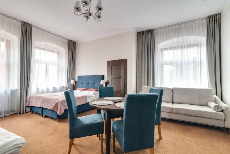 The bright room of Pensjonat Lider combines a bedroom and a seating area with a bed, a sofa, a table, turquoise chairs, and tall windows.