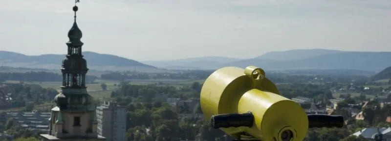 Depuis la forteresse de Kłodzko, on peut admirer un panorama de la ville avec le clocher de l'église, les toits rouges et une longue-vue jaune au premier plan, sur fond de montagnes douces.