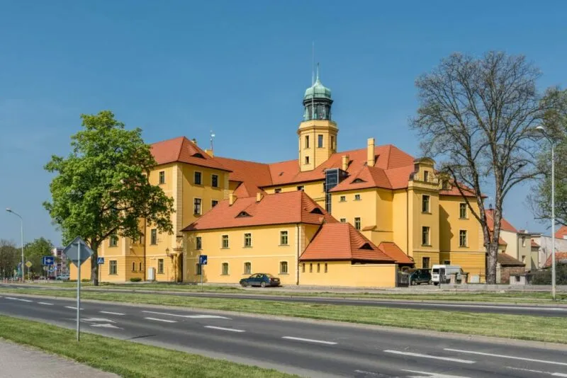 Ceglana, czerwonawo-brązowa budowla z jasnymi detalami wokół okien i bram, wyróżniająca się wieżą w kolorze czerwonej cegły.