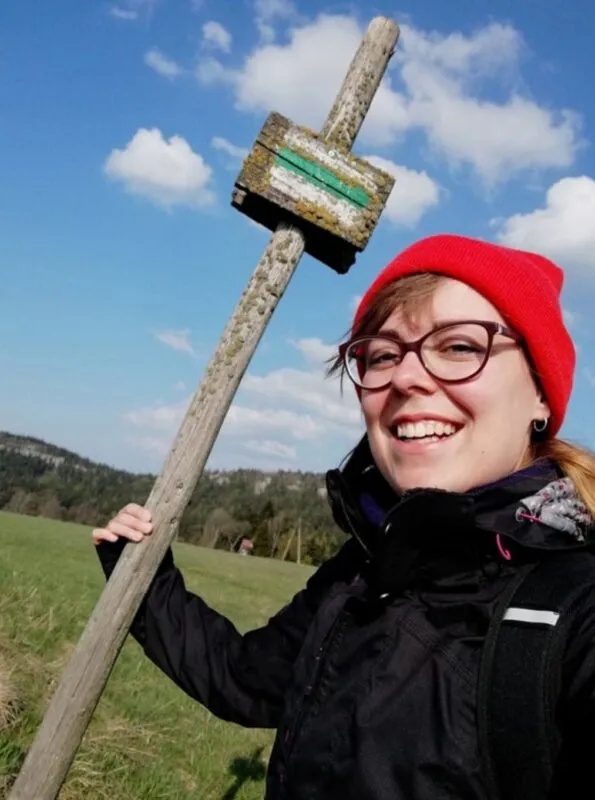 Kobieta w okularach, ubrana w czarną kurtkę oraz czerwoną czapkę, trzymająca drewniany znak oznaczający szlak
