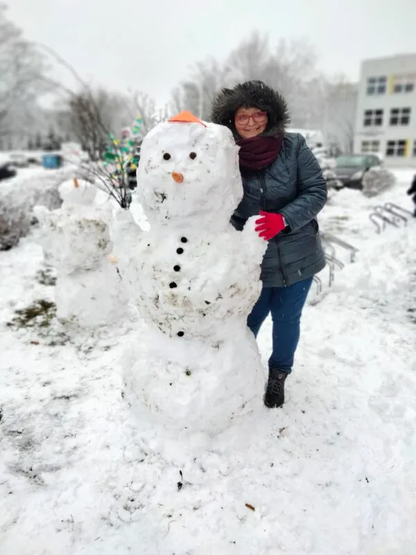 kobieta ubrana w ciepłą kurtkę oraz rękawiczki, stojąca razem ze zrobionym ze śniegu bałwanem, zdjęcia wykonane zimą, a na ziemi leży mnóstwo śniegu