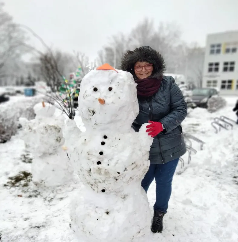 kobieta ubrana w ciepłą kurtkę oraz rękawiczki, stojąca razem ze zrobionym ze śniegu bałwanem, zdjęcia wykonane zimą, a na ziemi leży mnóstwo śniegu