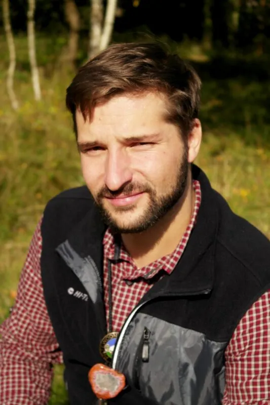 mężczyzna z brodą, w czerwonej koszuli i czarnej kamizelce, stojący na tle drzew