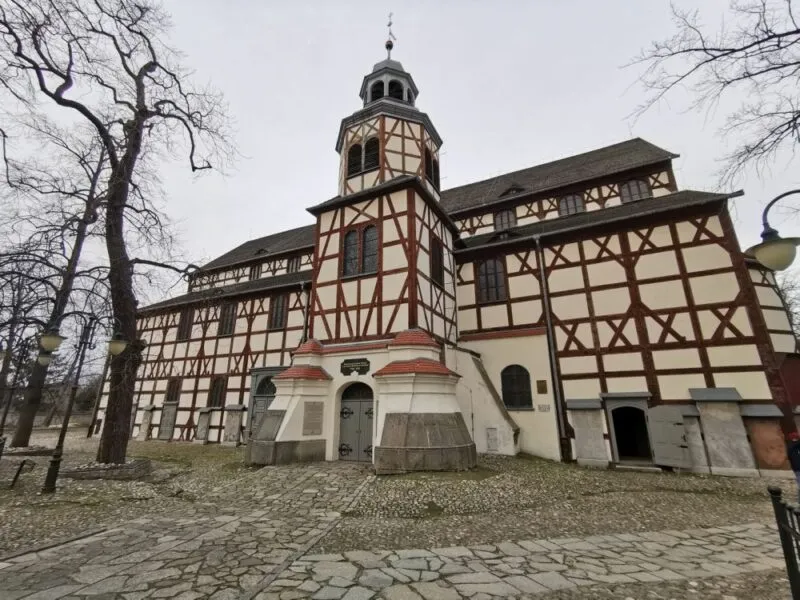 Na zdjęciu widoczny jest Kościół Pokoju w Jaworze, charakterystyczna budowla zbudowana z drewna, wpisana na listę UNESCO. Fasada kościoła wyróżnia się biało-brązowym układem drewnianych belek oraz centralną wieżą z barokowym hełmem. Otaczają go stare, bezlistne drzewa oraz brukowana ścieżka prowadząca do wejścia.