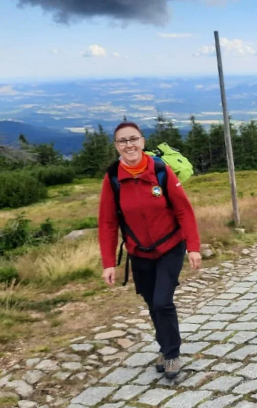 kobieta w czarnych spodniach i czerwonym polarze z plecakiem w kolorze jasno zielonym, na tle gór