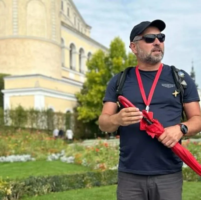 Mężczyzna trzymajacy czerwony parasol, stojący na tle dużego budynku o kolorze beżowym,