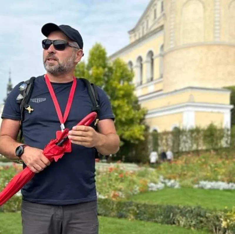 Mężczyzna trzymajacy czerwony parasol, stojący na tle dużego budynku o kolorze beżowym,