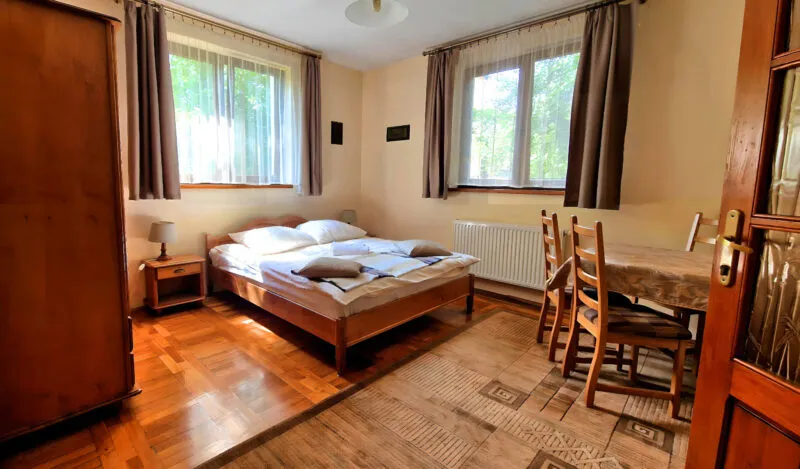 Wnętrze pokoju noclegowego, z podwójnym łóżkiem, szafą i stołem z krzesłami. 