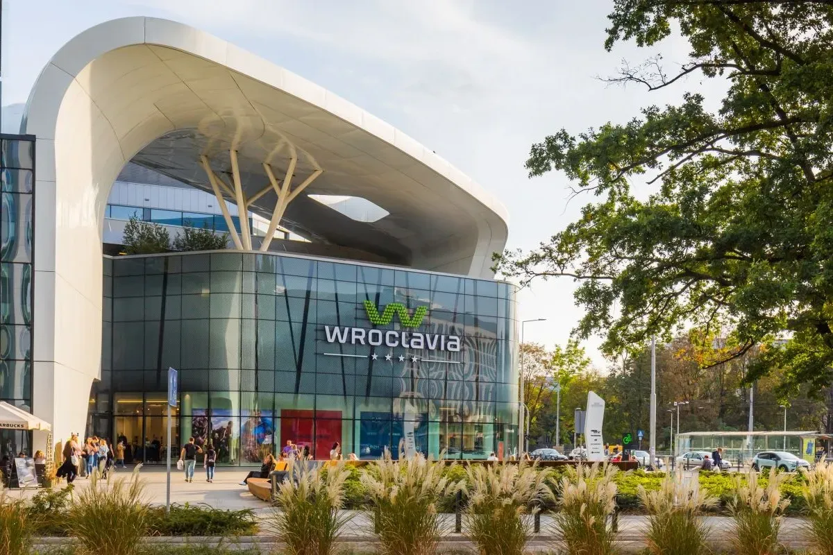 zdjęcie galerii handlowej Wroclavia o zachodzie słońca, sama galeria znajduje się na drugim planie a przed znajduję się pięć rownolegle ułożonych krzaków oraz po lewej jest wysokie drzewo. przy krzakach znajdują się zaokrąglone drewniane ławki na którzych siedzą ludzie. obok ławek idą ludzie z torbami na zakupy