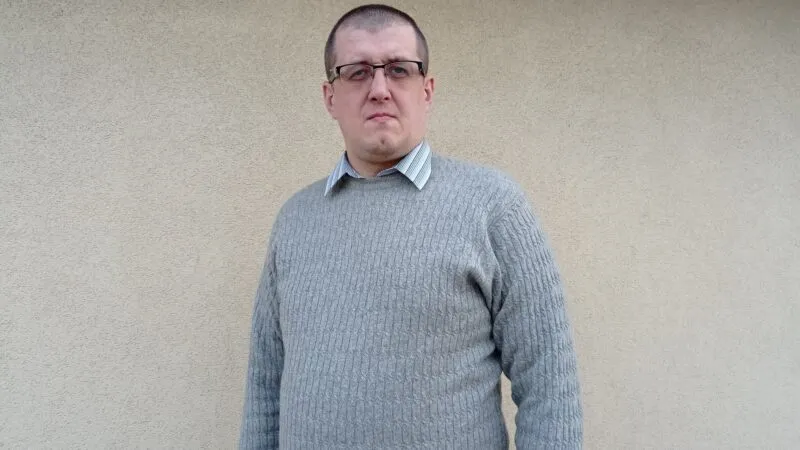 mężczyzna w okularach ubrany w szary sweter, stojący na tle szarej ściany