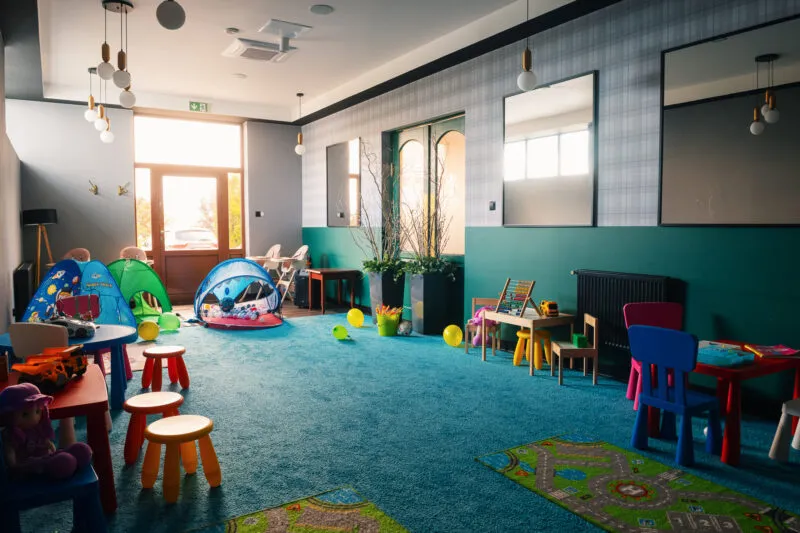 Duża przestronna sala zabaw, z dużą ilością stolików i krzesełek dziecięcych, małych namiotów oraz innych zabawek.