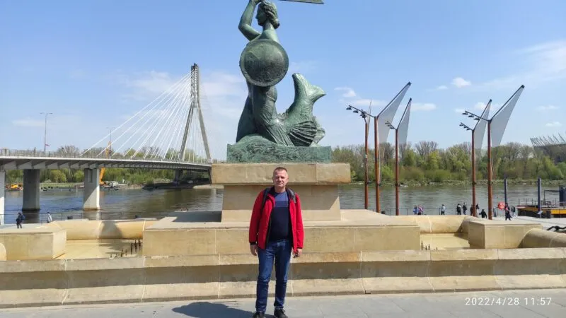 mężczyzna w czerwonym polarze, stojący na tle pomniku syrenki w Warszawie