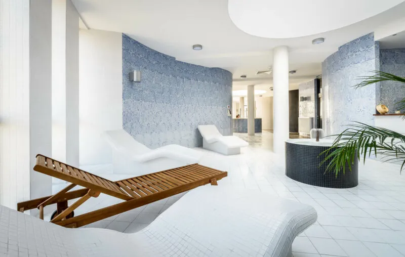 spa z drewnianymi oraz białymi, zaokrąglonymi leżakami. całośc jest wykonania estetycznie w białych kolorach. na dalekim planie znajduję się jacuzzi