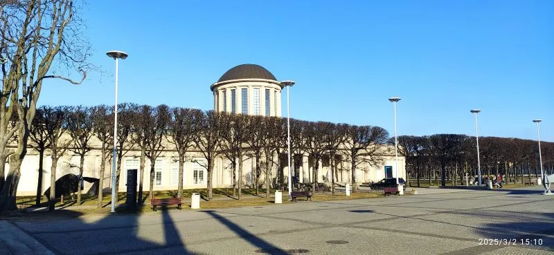 Modernistyczny budynek o prostych, geometrycznych kształtach, wyróżniający się czterema charakterystycznymi kopułami na dachu, otoczony przestronnym terenem.