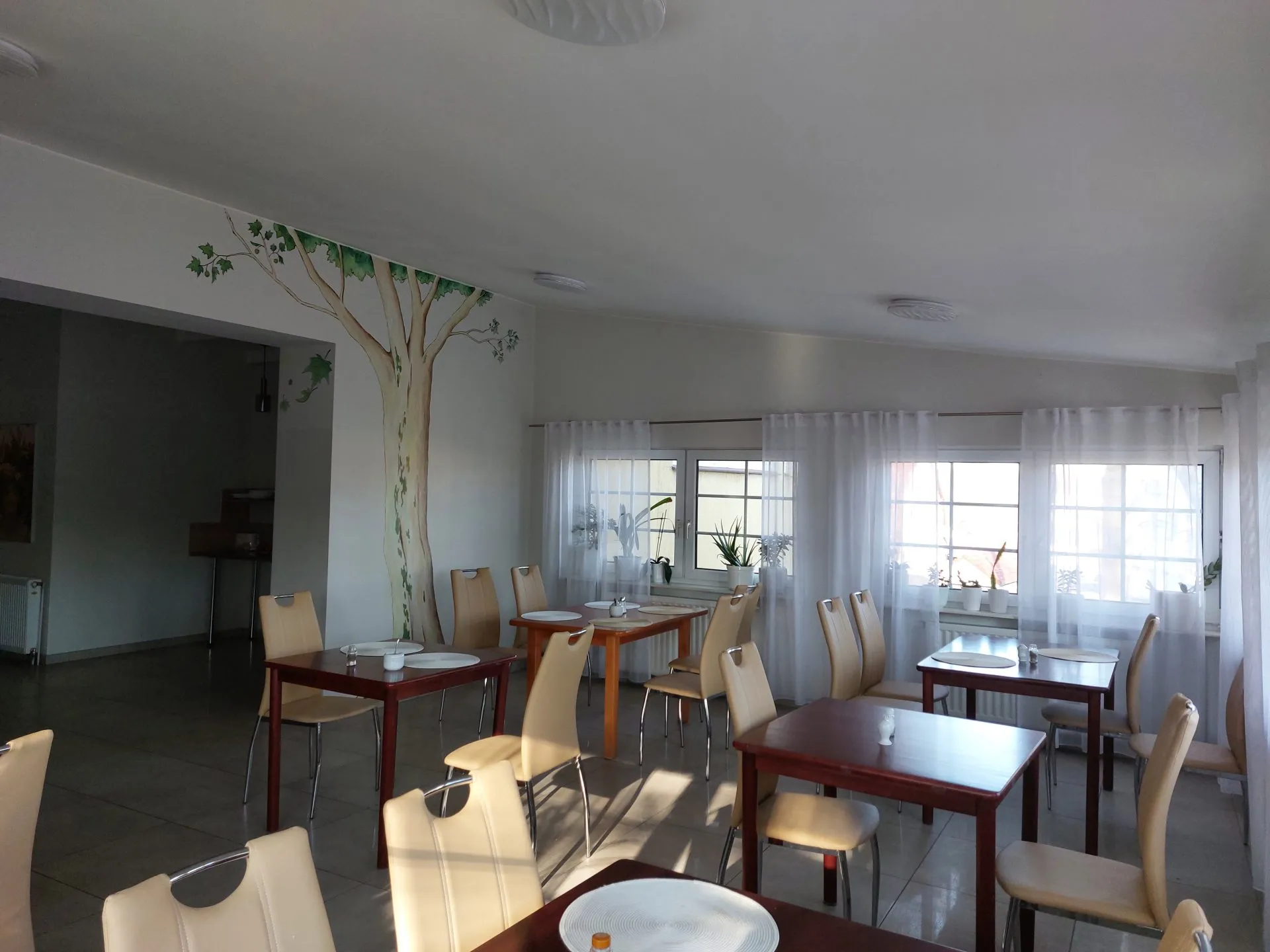 Sala restauracyjna z kilkunastoma stolikami oraz krzesłami. Na podłodze płytki, na jednej ze ścian namalowany platan.