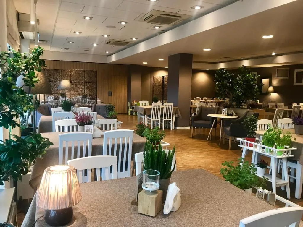 Przestronne, eleganckie wnętrze restauracji z białymi drewnianymi krzesłami oraz prostokątnymi stołami nakrytymi obrusami. Dekorację stanowią małe lampki, dużo roślin zielonych oraz świeczki.