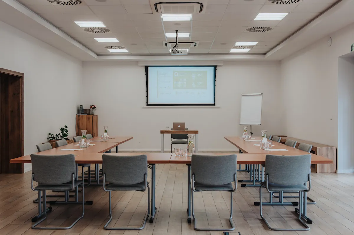 Sala konferencyjna z podłużnymi stołami ułożonymi w literę U i przylegającym doń krzesłami. Na ścianie ekran multimedialny.