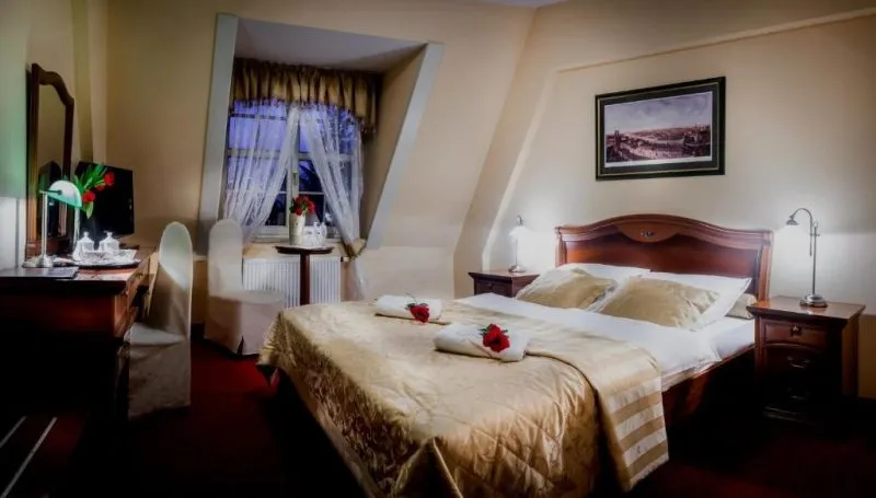 Elegancki, schludny pokój hotelowy z podwójnym łożem małżeńskim, komodą oraz krzesłem, dwoma szafkami nocnymi z lampkami i małym stoliczkiem.