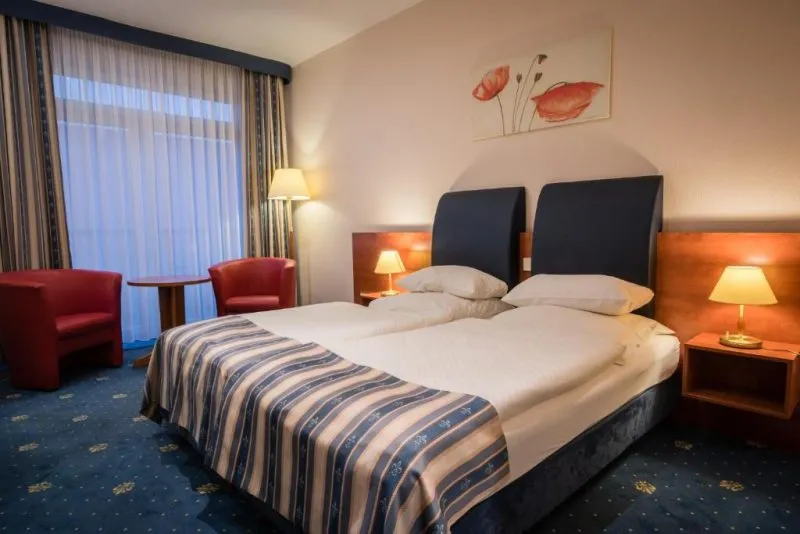 Pokój hotelowy z dużym łóżkiem podwójnym, szafkami nocnym i lampkami. Pod oknem okrągły stolik i dwa czerwone fotele. Na ścianie nad łóżkiem namalowane maki.