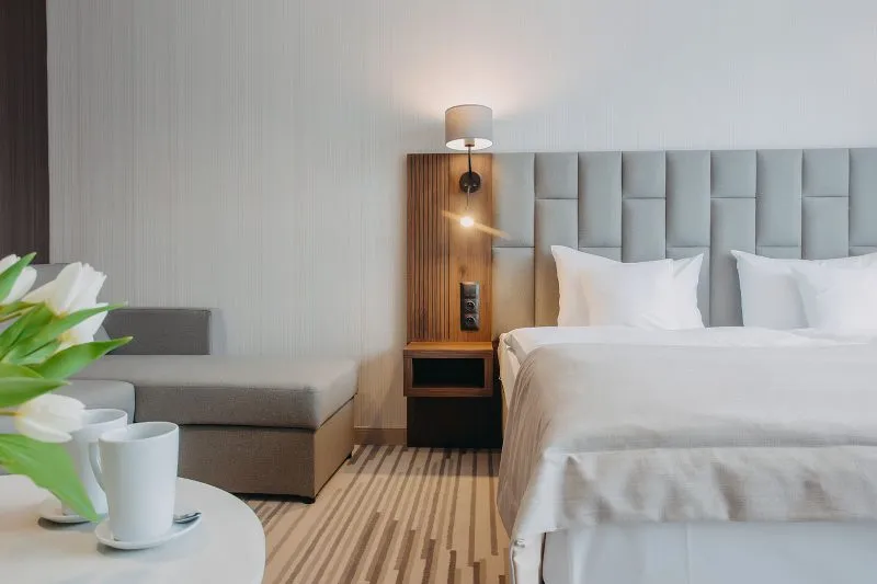 Nowoczesny, pokój hotelowy z podwójnym łóżkiem z zagłówkiem obitym tkaniną. Na podłodze wykładzina. Widoczny kawałek kolistego stolika i sofy.