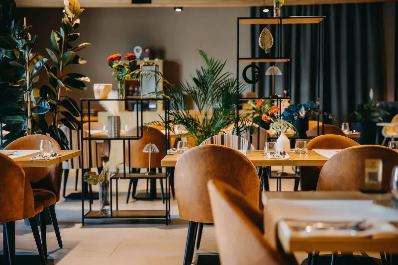 Nowoczesna, designerska restauracja hotelowa z elementami drewnianymi oraz metalowymi. Dekorację stanowią rośliny zielone, długie zasłony, drobne przedmioty ozdobne.