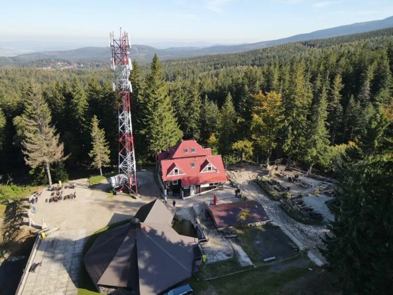 Zdjęcie z drona przedstawiające rozległy plac w lesie iglastym, na terenie którego ustawiony jest budynek schroniska, duża wiata oraz maszt nadajnikowy.