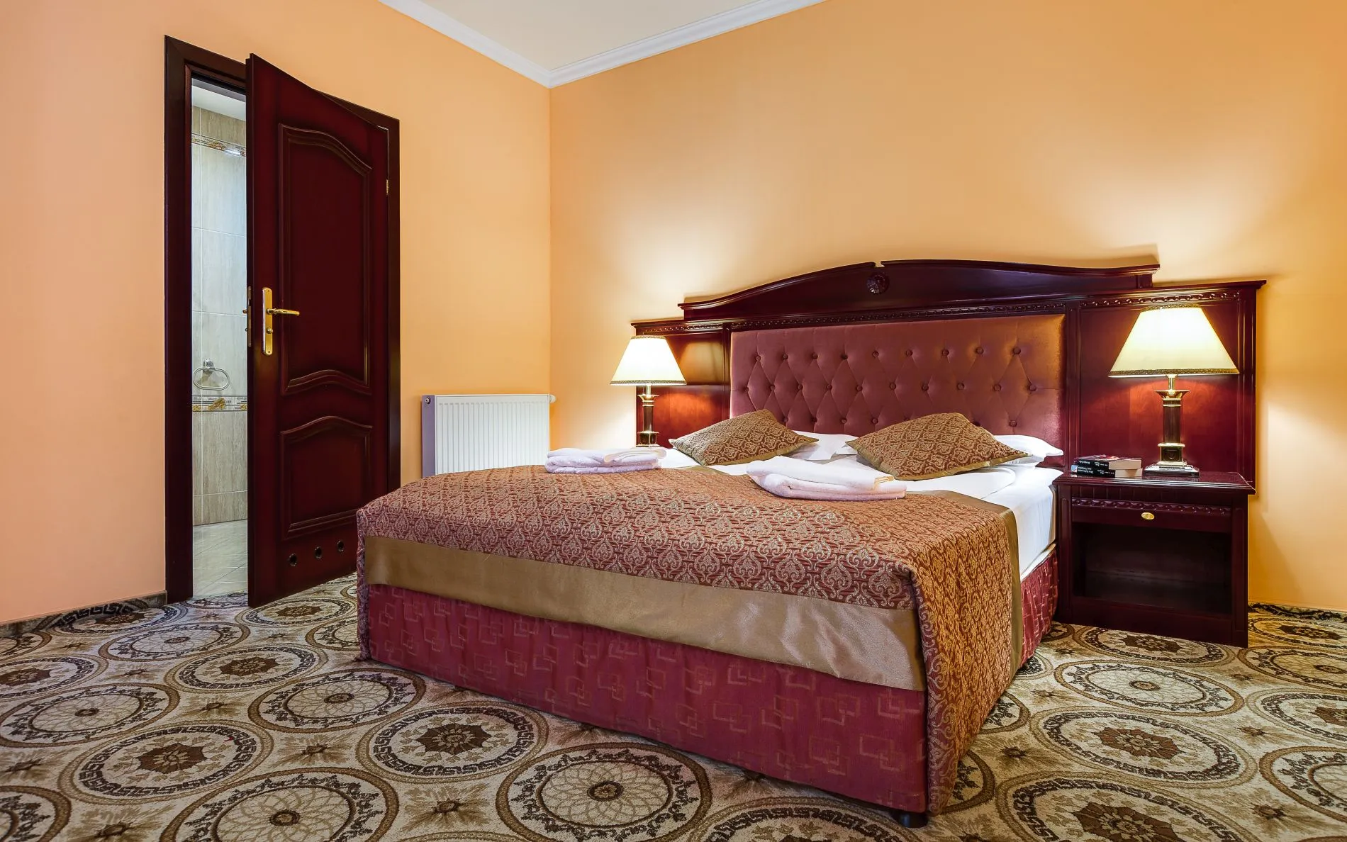 Pokój hotelowy z dużym podwójnym łóżkiem. Po bokach szafki nocne z lampkami. Na podłodze wykładzina.