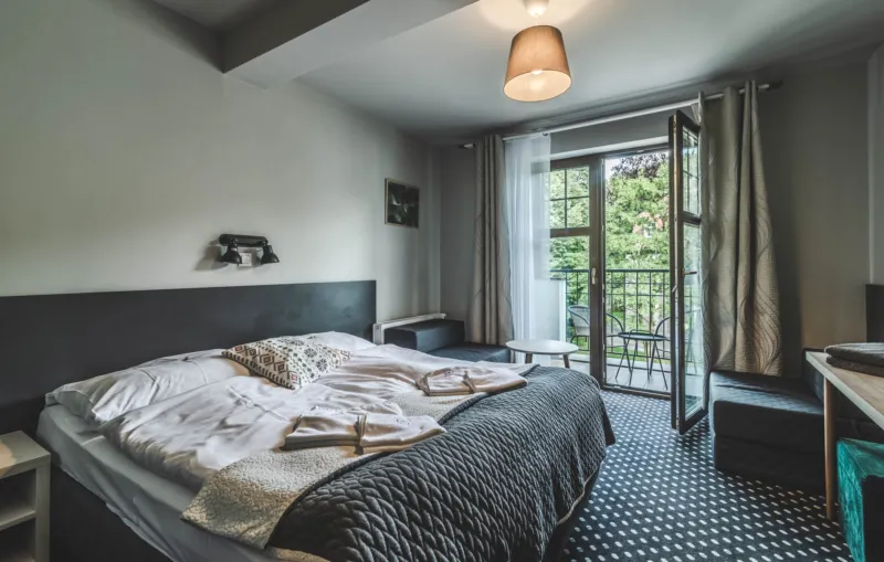 Pokój hotel z podwójnym łóżkiem. Szerokie okno prowadzi na balkon.