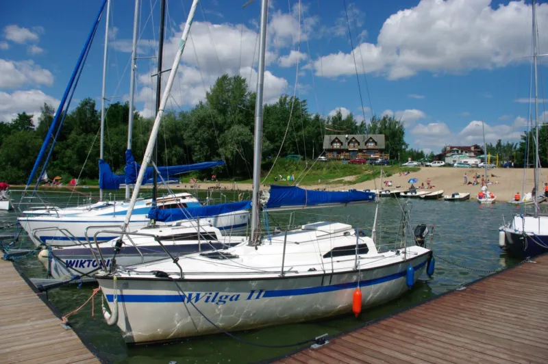 Widok na łódki przy pomoście, w tle plaża i drzewa.