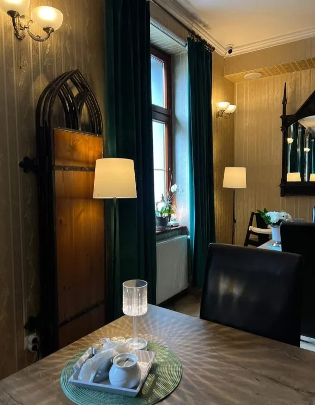 Lobby hotelowe. Stoły z czarnymi fotelami, stojące lampy oraz lustro w drewnianej ramie.