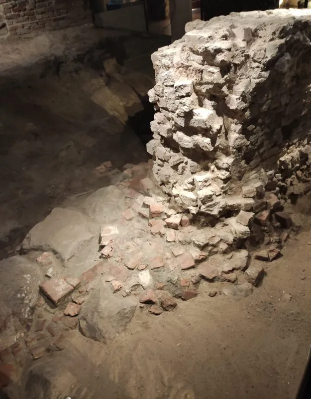 Ruiny z cegieł i kamieni, częściowo zasypane piaskiem. To pozostałości dawnej konstrukcji odsłonięte podczas badań archeologicznych.