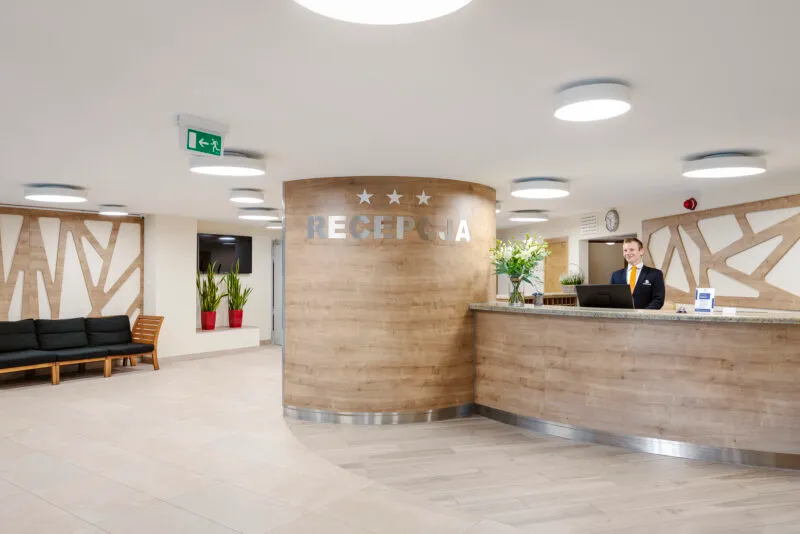 Elegancka recepcja hotelowa w odcieniach jasnego drewna i bieli. Lada przylega do szerokiej kolumny.