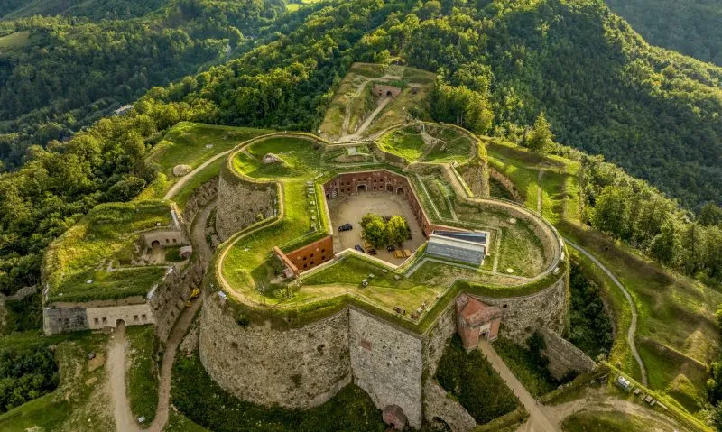 Majestatyczna twierdza górska na tle gór porośniętych gęstymi zielonymi lasami.
