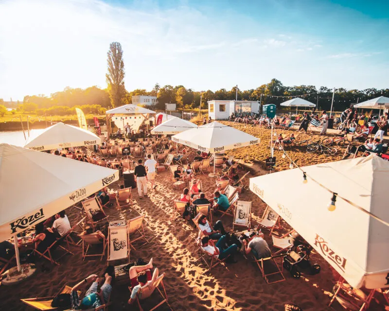 Zdjęcie wykonane przy słonecznej, letniej pogodzie, przedstawia bar na plaży z gośćmi siedzącymi na leżakach.