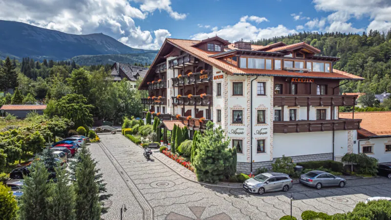 Budynek hotelu w stylu tyrolskim na tle lasu oraz gór.