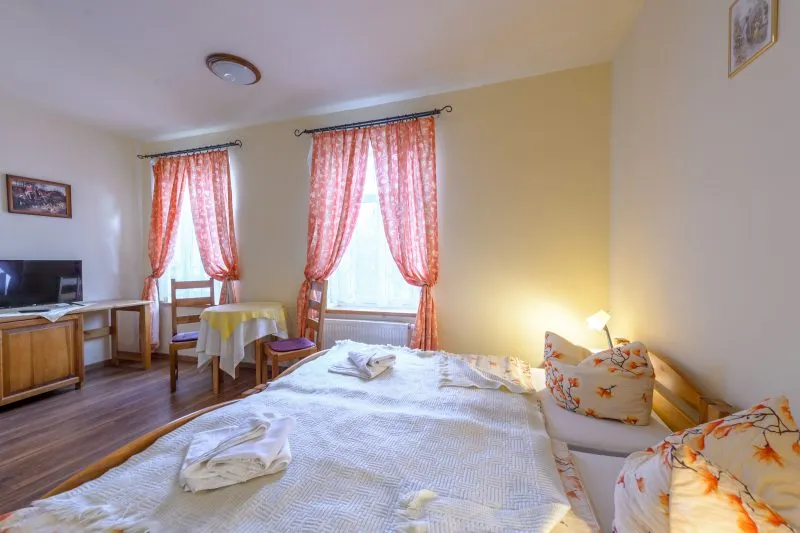 Pokój hotelowy z dwoma pojedynczymi łóżkami. Po przeciwległej stronie komoda z telewizorem. Pod oknem, przesłoniętym podpiętymi zasłonami, stolik z dwoma krzesłami.