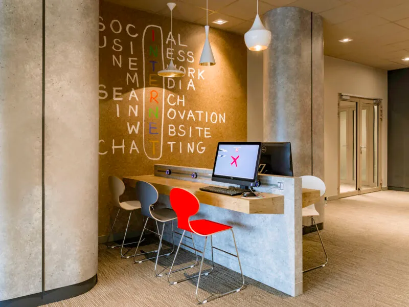 Lobby hotelowe w nowoczesnym modernistycznym stylu, ze stołem barowym z komputerami oraz nowoczesnymi krzesłami. Na ścianie modernistyczna fototapeta.