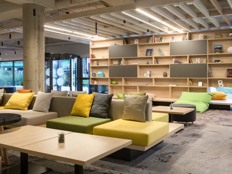 Hotelowe lobby z kanapami wypoczynkowymi oraz regałem z książkami i drobnymi elementami dekoracyjnymi.