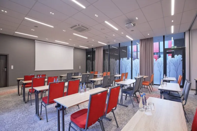 Sala konferencyjna z 4 rzędami prostokątnych stołów. Na ścianie duży ekran.