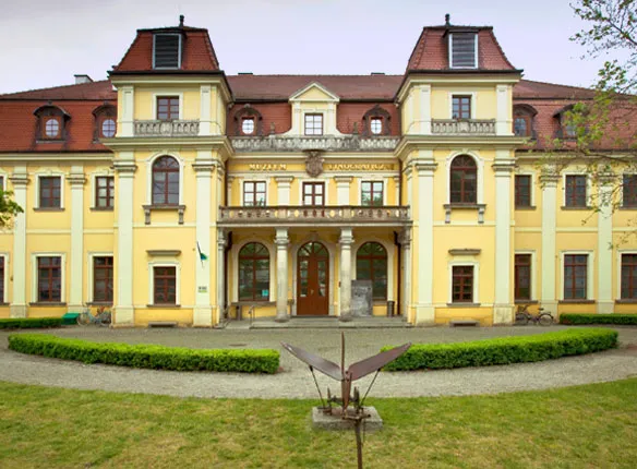 zdjęcie muzeum etnograficznego w lekko pochmurny dzień, przed obiektem znajdujęsię trawnik oraz żywopłot, budynek jest w kolorach zółto-białych, ma wiele okien, a dach jest skośny w kolorze ciemno czerwonym