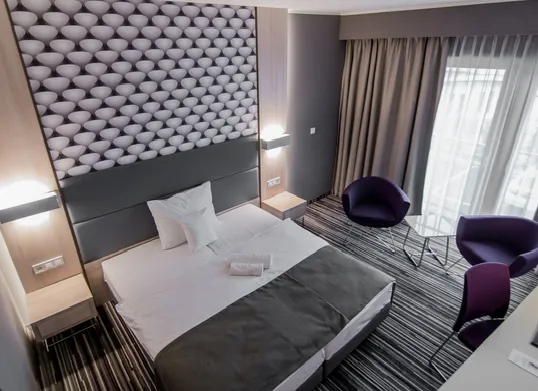 Zdjęcie przedstawia nowoczesny, elegancki pokój hotelowy jednoosobowy urządzony w minimalistycznej stylistyce. Dominują w nim biel i szarość. W pokoju znajdują się dwa pojedyncze łóżka ustawione obok siebie. Na łóżku leży szara narzuta oraz ręczniki. W oknach długie ciężkie, beżowe zasłony.