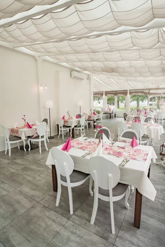 Pomieszczenie tarasowe zaadaptowane na restaurację. Stoły nakryte białymi obrusami i koliste krzesła. Delikatne różowe akcenty w postaci serwetek i bieżników.