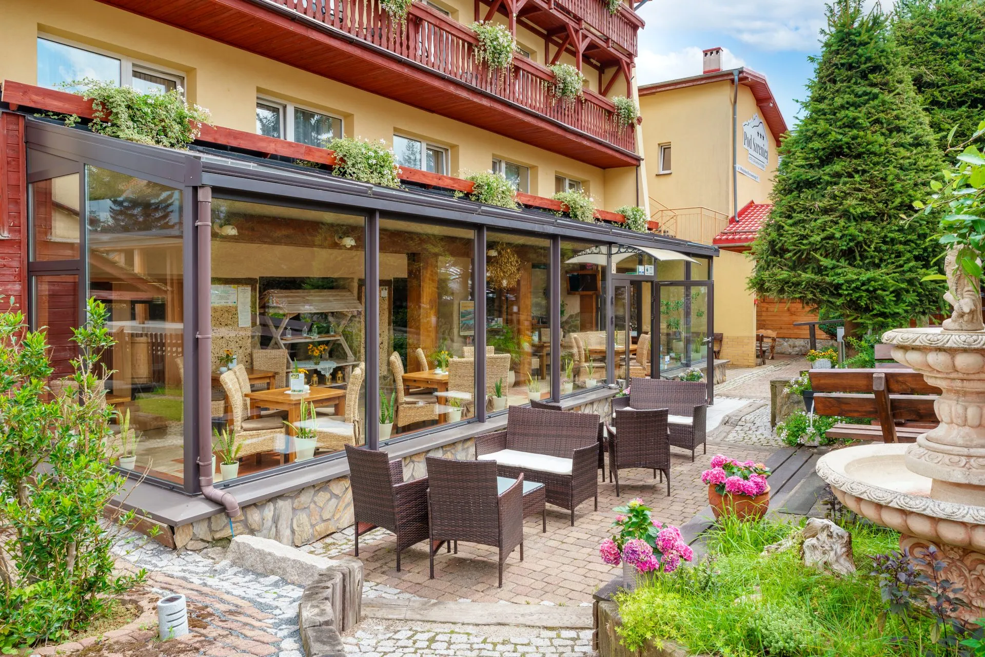 Przytulne patio hotelowe, z fontanną oraz zielenią ogrodową. Na brukowanej powierzchni ustawiono fotele i stoły ogrodowe. Do budynku przylega szklana weranda, gdzie znajdują się stoliki i krzesła restauracyjne.