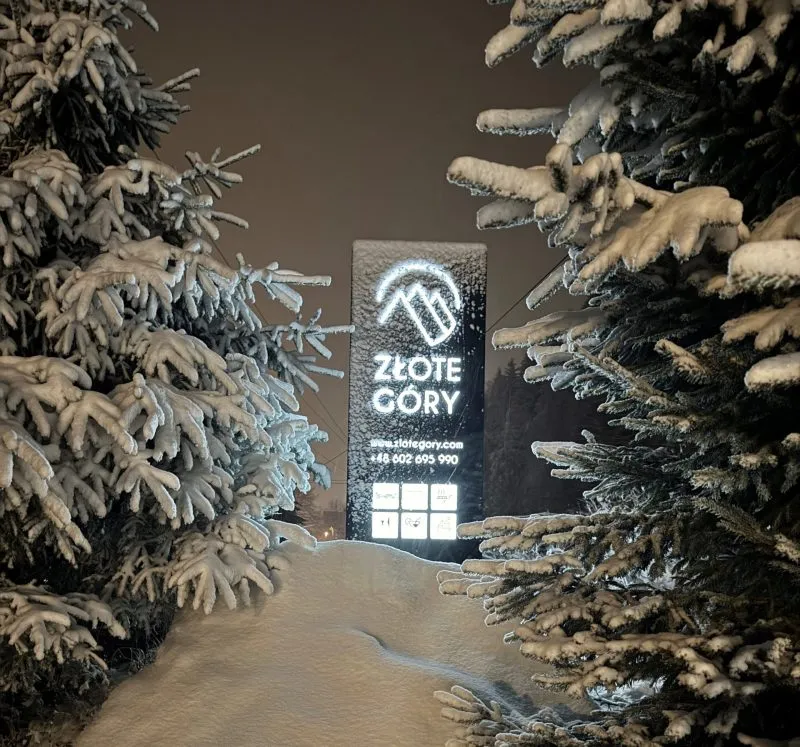 Zimowy krajobraz z dużą ilością śniegu, otaczającego podświetlony znak hotelu "Złote Góry". Światło reklamy kontrastuje z ciemnością nocy, tworząc malowniczy widok wśród ośnieżonych drzew.