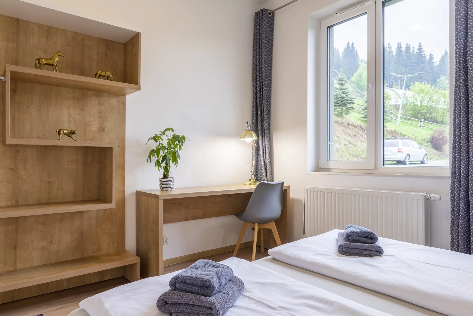 Nowoczesny pokój hotelowy z dwoma łóżkami, biurkiem oraz dużym oknem, przez które rozpościera się widok na górski krajobraz. Wnętrze jest utrzymane w naturalnych, ciepłych barwach, z drewnianymi elementami i minimalistycznymi dekoracjami, takimi jak złote figurki zwierząt na półkach.