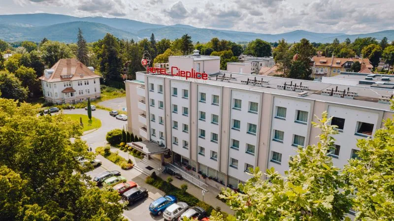Duży prostokątny budynek z napisem "Hotel Cieplice" na szczycie fasady, na tle zieleni oraz gór.