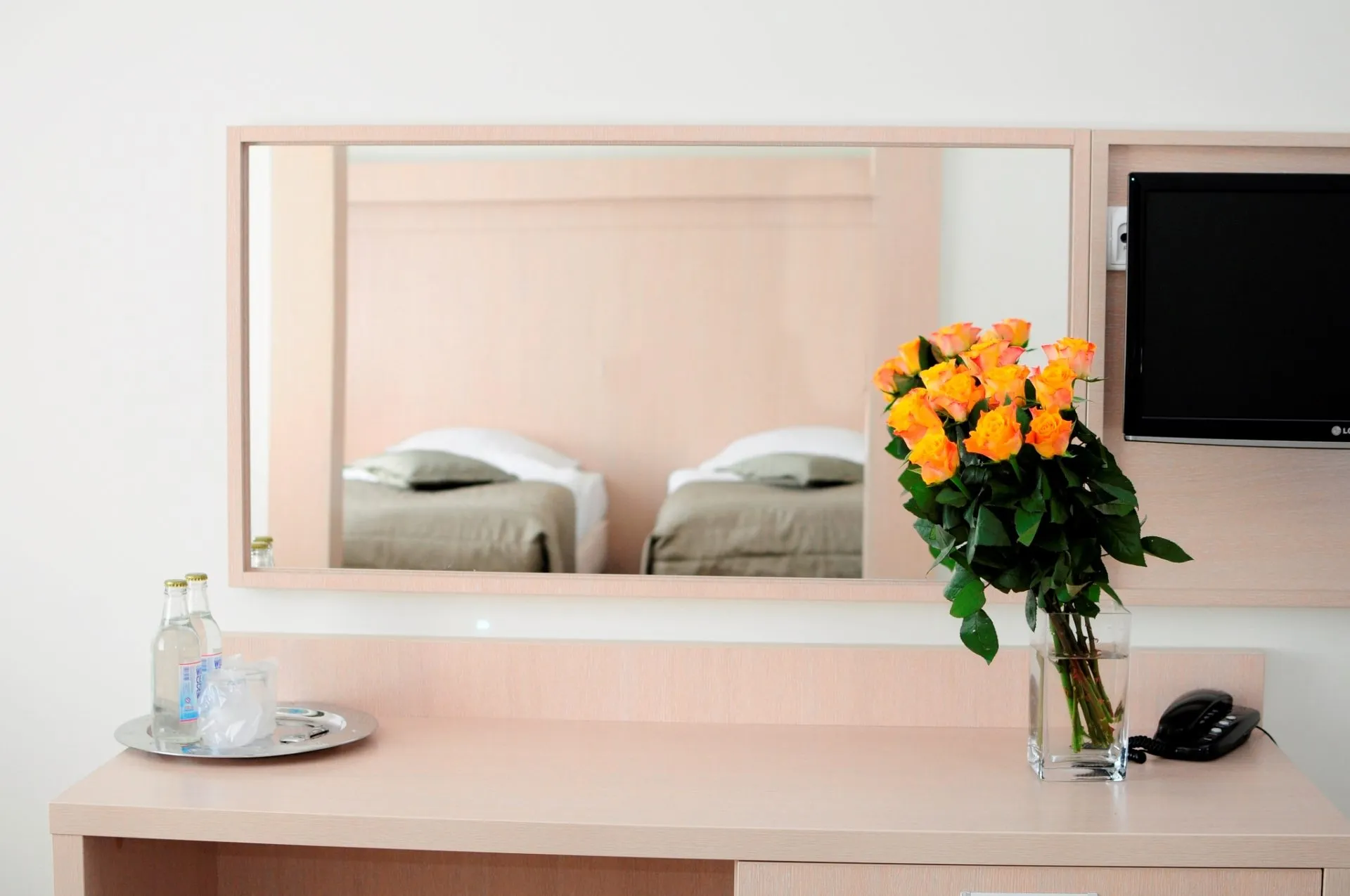 Lustro w pokoju hotelowym z odbiciem dwóch łózek pojedynczych. Pod lustrem blat z wazonem z pomarańczowymi różami oraz taca z wodą mineralną.