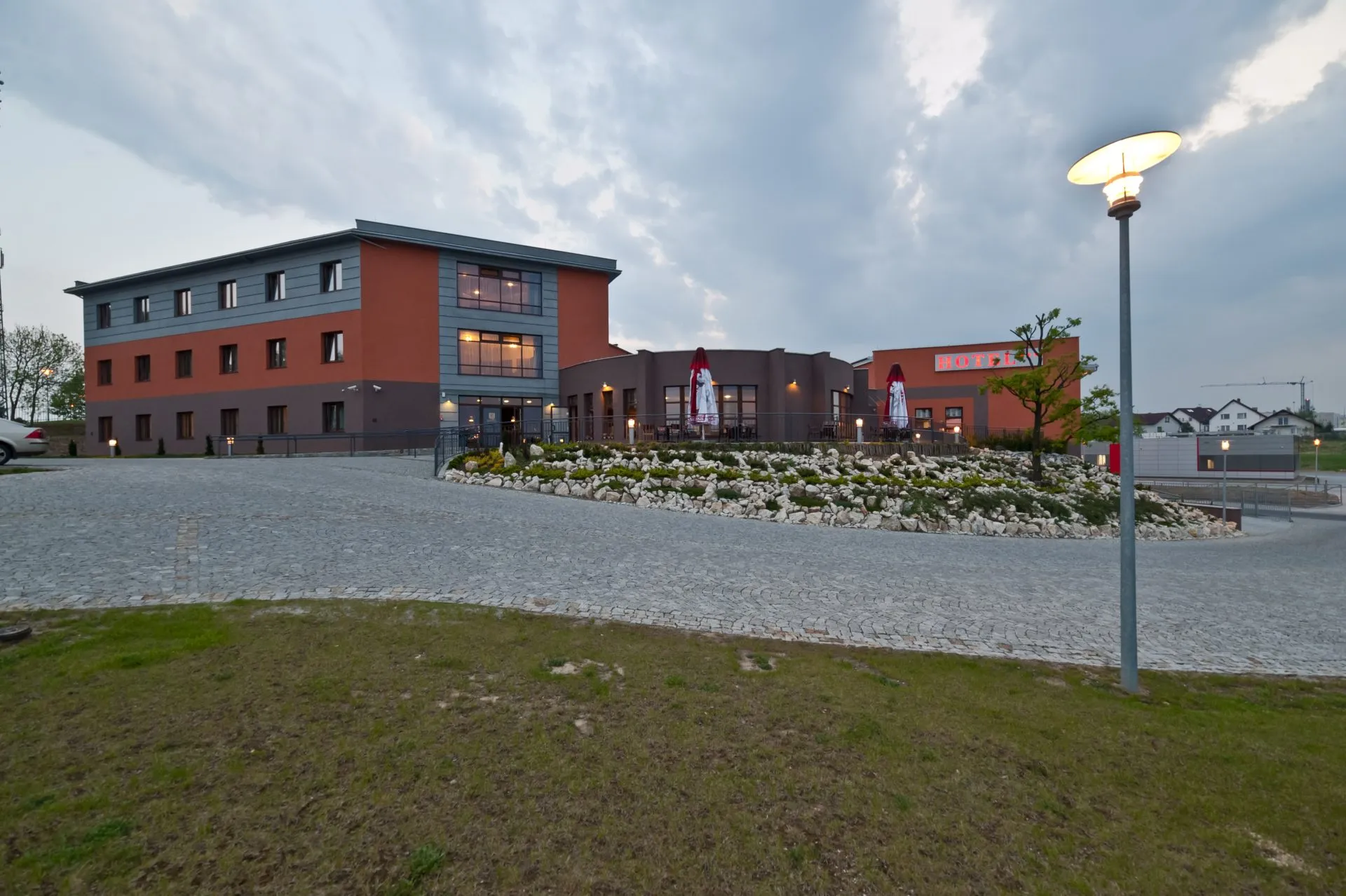 Widok na Hotel Esperanto w Świdnicy, nowoczesny budynek o ceglano-grafitowej elewacji, otoczony elegancko zagospodarowanym terenem. Podjazd wyłożony kostką brukową prowadzi do głównego wejścia, a oświetlenie i ogródek z kamieniami dodają mu przytulnego charakteru.